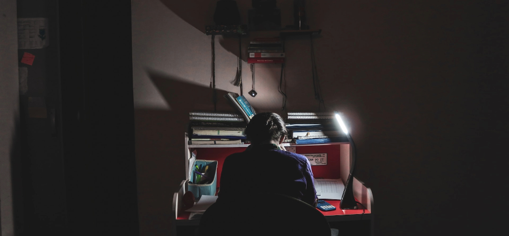 Man with his back to the camera sitting at a desk in a dark room,