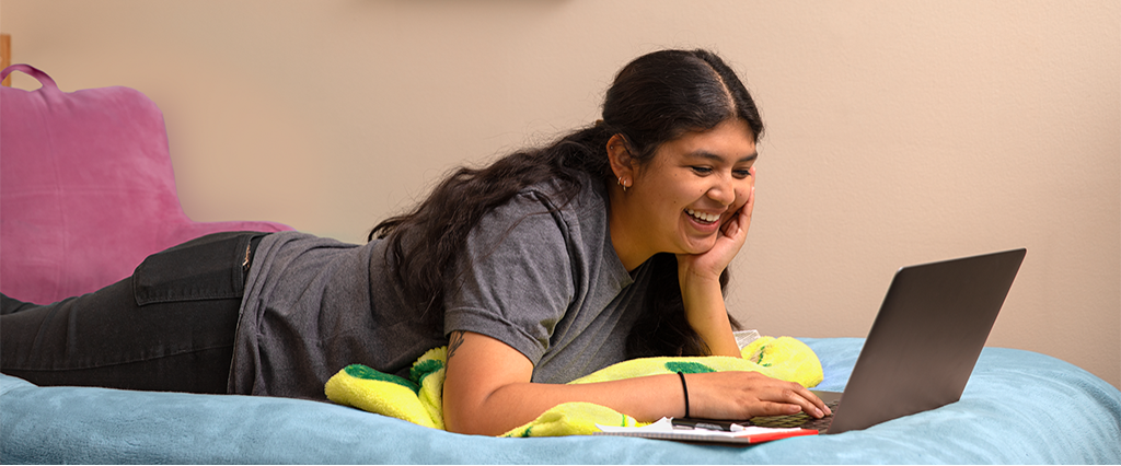 College student laying on a bed using a laptop computer.