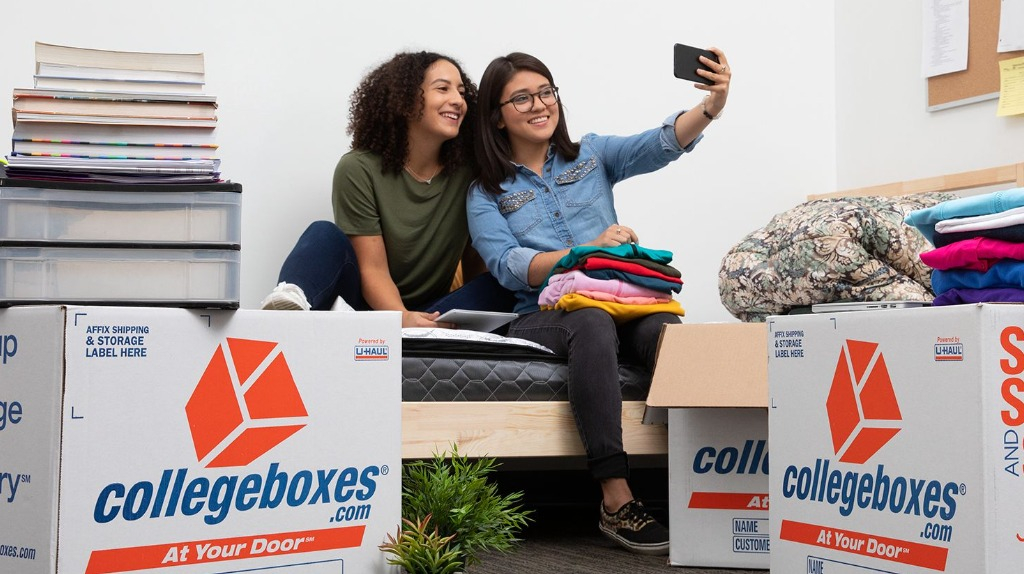 Two young women in a dorm room sit on a bed and take a selfie, surrounded by moving boxes and textbooks.