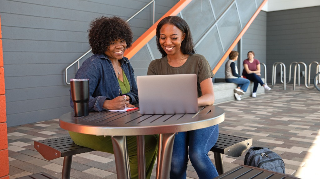Two African American women sitting at an outdoor table on a college campus. They are both smiling and looking at a laptop screen.
