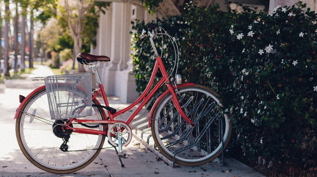 A lone red bicycle parked outside at a bike rack in front of a bush.