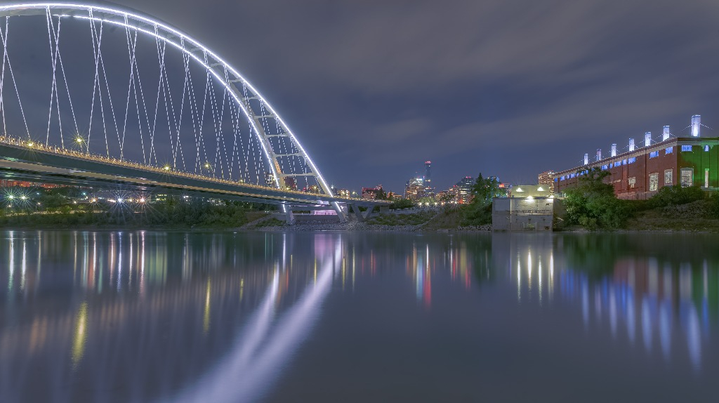 Nighttime scene of a bridge against a city landscape. The lights of the city and bridge glow in the water.