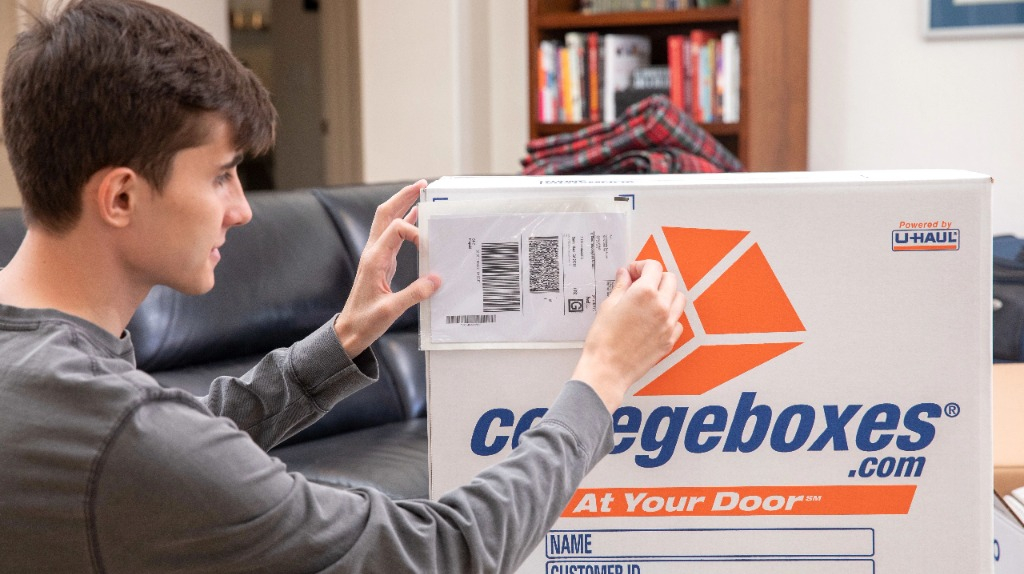Young brunette man placing a shipping label on a Collegeboxes box.