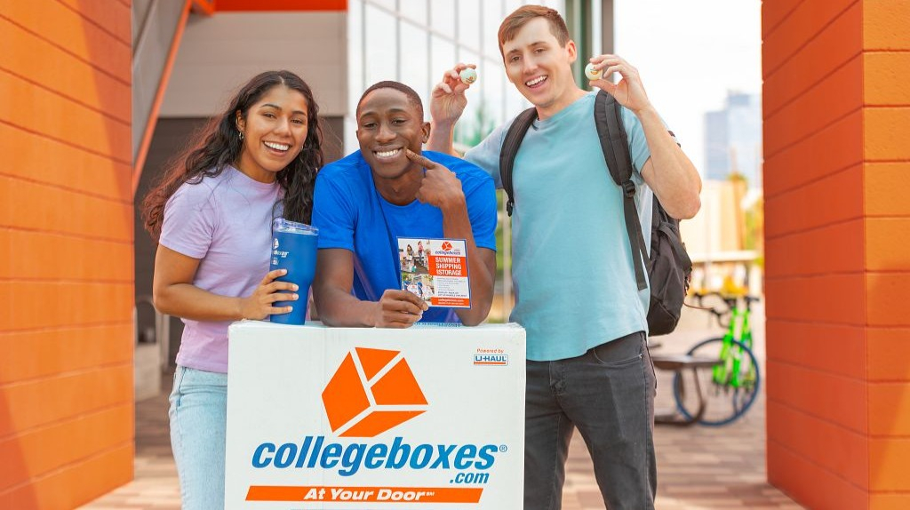 3 students gather around a Collegeboxes box and smile.