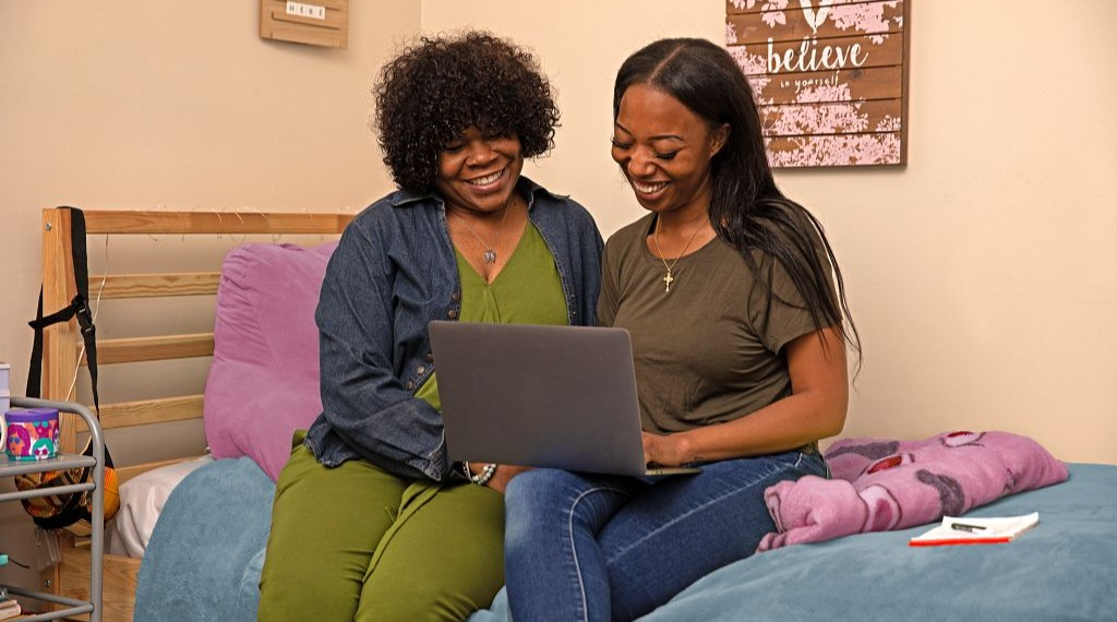 Two women sitting on a bed and looking at a laptop on the younger woman's lap.