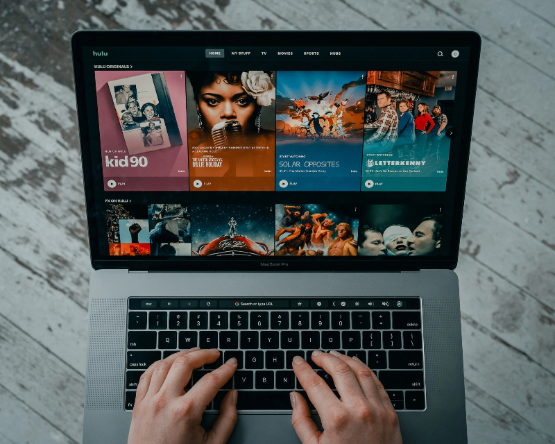 A pair of hands hovering over a laptop displaying a Hulu homepage.