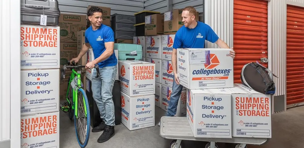 Two men in blue shirts moving boxes and a bike from a storage unit.