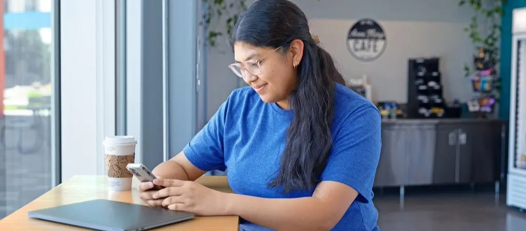 Girl sitting at a table on her phone with a laptop and coffee