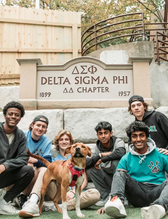 6 boys and a dog posing in front of Delta Sigma Phi