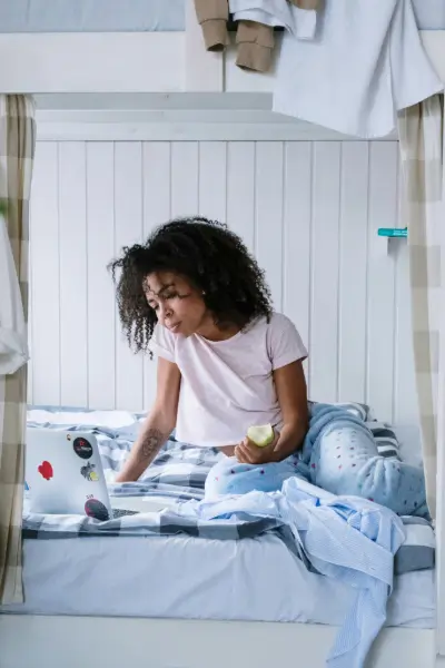 Girl working on her bed with a computer, eating an apple