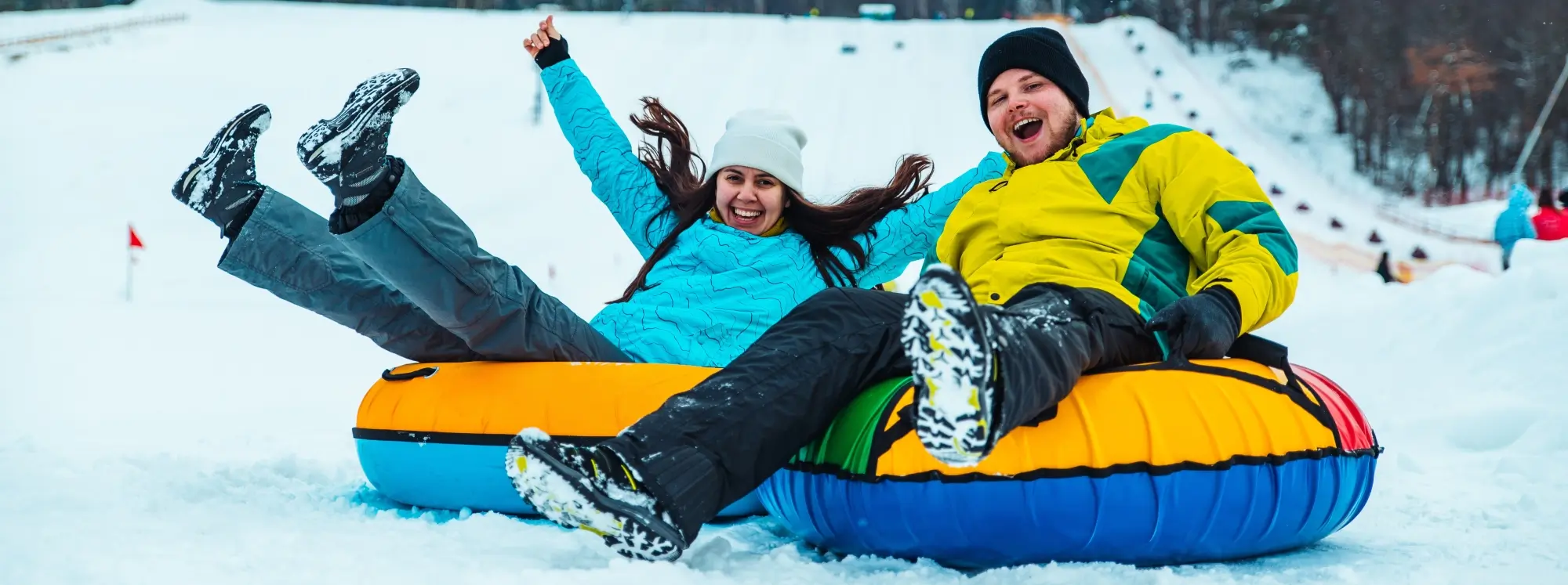 Two people joyfully snow tubing down a hill, wearing colorful winter jackets and hats, with their feet up in the air, amidst a snowy landscape.